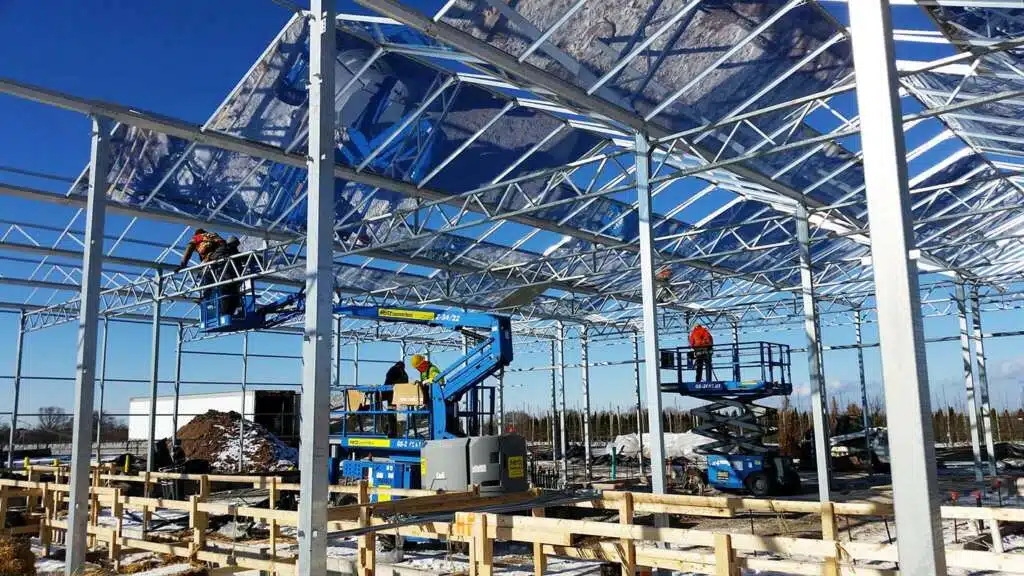 Workers on lifts assembling the steel frame and roof panels of a large commercial greenhouse under construction.