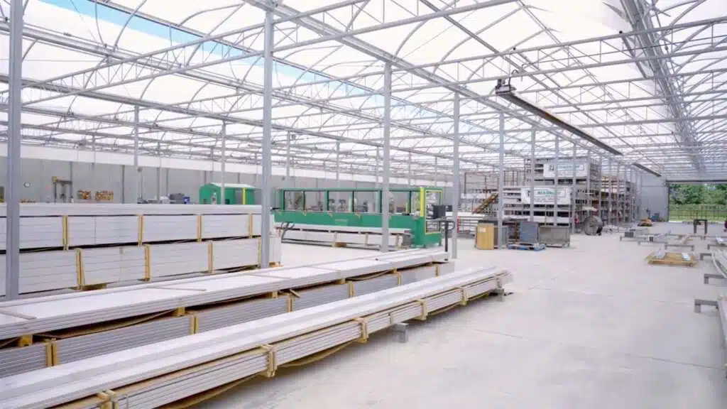 Interior of a large industrial warehouse, possibly a greenhouse manufacturing facility, with stacks of building materials and machinery.